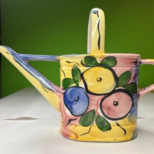 Puede incluir: Una regadera de cerámica pintada a mano con base amarilla, adornada con diseños florales azules, rosas y amarillos y hojas verdes. El pico y el asa son azules y amarillos. Tiene un asa vertical.