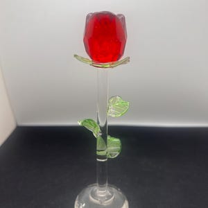 Puede incluir: Una escultura de rosa de cristal transparente con una flor de rosa roja facetada, hojas verdes y un tallo transparente. La rosa mide aproximadamente 15 cm de altura y se apoya sobre una base redonda. El cristal es transparente.