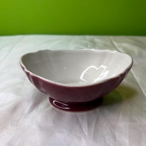 Może przedstawiać: Mała, dekoracyjna miska z falistym brzegiem. Zewnętrzna strona jest w kolorze głębokiego bordowego, a wewnętrzna biała. Misa stoi na małej, okrągłej podstawie.