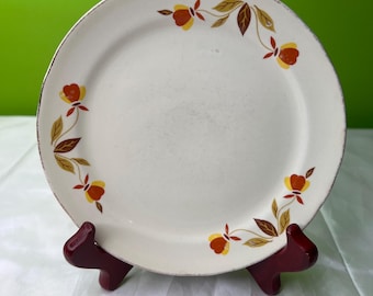 Puede incluir: Un plato de cerámica blanco con un diseño floral con flores naranjas y amarillas con hojas marrones. El plato tiene un borde dorado y se exhibe en un soporte rojo oscuro.