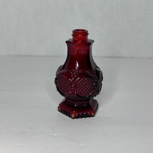 Puede incluir: Una botella de vidrio rojo oscuro con un diseño decorativo. La botella tiene un cuerpo redondo y una base hexagonal.
