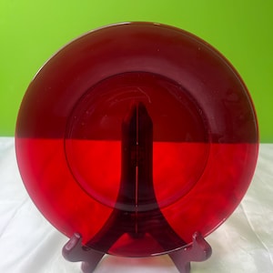May include: A vibrant red glass plate displayed on a dark brown stand. The plate is circular with a glossy finish, reflecting light. The stand has a three-legged design, providing support. The background is a solid green.