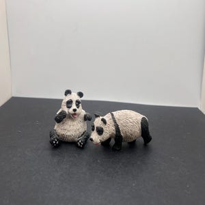 May include: Two panda figurines. One panda is sitting with its tongue out, and the other is walking. Both pandas have black and white fur, with black markings around their eyes, ears, and limbs.