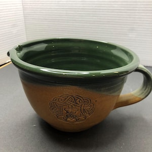Puede incluir: Una taza de cerámica marrón con un esmalte verde. La taza tiene un asa y un diseño de nudo celta con un trébol en el centro.