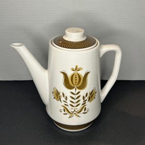 Op de afbeelding: Witte koffiekan van keramiek met een bruin bloemenmotief. De kan heeft een deksel met een bruin stippenpatroon.