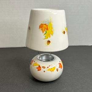 Pode incluir: Aquecedor de velas de cerâmica branca com um abajur branco. O abajur tem um design de folhas de outono em tons de laranja, amarelo e marrom. A base do aquecedor tem um design de folhas de outono e bolotas.