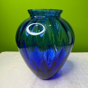 May include: A decorative glass vase with a rounded body and a ruffled rim. The vase features a blend of blue and green hues, with darker shades at the base. The glass has a glossy finish, reflecting light.