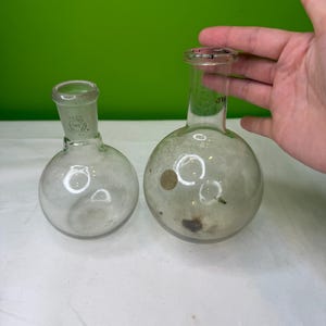 Może przedstawiać: Dwa przezroczyste kolby okrągłodenne z wąskimi szyjkami. Na jednej kolbie znajduje się etykieta z napisem "PYREX". Kolby są wykonane ze szkła i prawdopodobnie służą do eksperymentów naukowych lub prac laboratoryjnych.