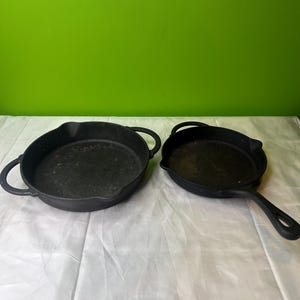 May include: Two black cast iron skillets. One skillet has two loop handles, and the other has a single long handle. Both skillets are round with a slightly raised edge. The background is a solid green.