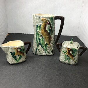 Op de afbeelding: Een vintage keramische set met een roomkan, suikerpot en kan met een grijs en groene glazuur. Elk stuk heeft een gestileerd hertontwerp in bruin en wit.