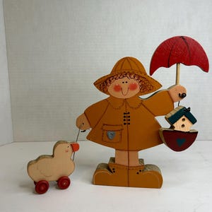 Può includere: Una figurina di legno di una ragazza che indossa un impermeabile giallo e un cappello rosso, tiene un ombrello rosso e una casetta per gli uccelli. Sta passeggiando un piccolo anatroccolo di legno su ruote.