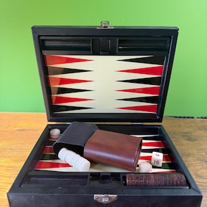 Puede incluir: Juego de backgammon negro abierto con tablero rojo, negro y blanco. Incluye cubiletes marrones, fichas de madera y un dado blanco que muestra el número 16. El juego está sobre una superficie de madera.