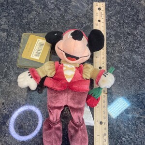 May include: A plush Mickey Mouse doll dressed in a red velvet vest and pants with tan sleeves, holding a red rose. A wooden ruler placed next to the doll shows a height of approximately 8 inches. A gold tag and a purple hair tie are also visible.