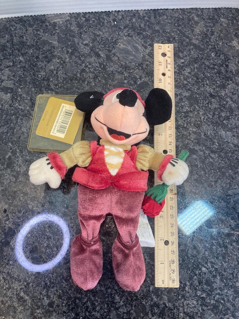 May include: A plush Mickey Mouse doll dressed in a red velvet vest and pants with tan sleeves, holding a red rose. A wooden ruler placed next to the doll shows a height of approximately 8 inches. A gold tag and a purple hair tie are also visible.
