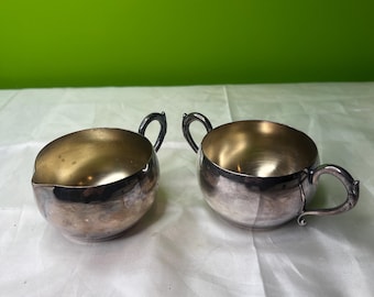 Puede incluir: Dos azucareros y cremeros antiguos plateados. Los cuencos tienen forma redondeada con exterior plateado brillante e interior dorado. Cada cuenco tiene un asa pequeña y curvada. El fondo es una superficie clara con un fondo verde.