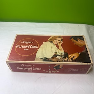 May include: A vintage Scrabble Crossword Cubes game box. The box has a red and brown color scheme with the game title in white. The box features an illustration of a man and woman playing the game. The game is published by Selchow & Righter Co.