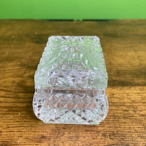 May include: A clear glass trinket box featuring a faceted diamond-cut pattern across its rectangular body and lid. The container has a slightly curved base and a geometric, textured surface that reflects light through its transparent material.