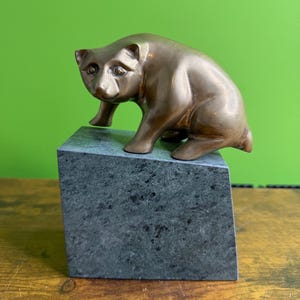 May include: A metallic bronze-toned bear figurine in a crouching position, mounted on top of a rectangular, dark gray, speckled stone base. The bear has smooth, polished surfaces and is depicted with its head turned slightly to the left.