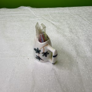 May include: A small, white ceramic mug with a handle, decorated with green leaf and vine designs. The mug contains a clear plastic wrapper filled with colorful items. The handle has blue stripes.