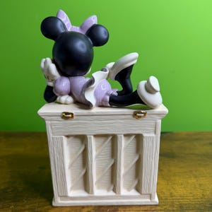 Peut inclure: Une figurine en c&eacute;ramique repr&eacute;sentant un personnage aux oreilles noires avec un n&oelig;ud et une robe violets, allong&eacute; sur une base rectangulaire couleur cr&egrave;me &agrave; texture bois&eacute;e. La base comporte deux petites charni&egrave;res en m&eacute;tal dor&eacute; sur le dessus.