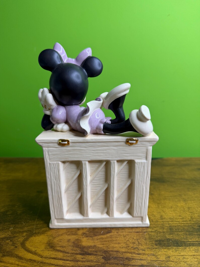 Peut inclure: Une figurine en c&eacute;ramique repr&eacute;sentant un personnage aux oreilles noires avec un n&oelig;ud et une robe violets, allong&eacute; sur une base rectangulaire couleur cr&egrave;me &agrave; texture bois&eacute;e. La base comporte deux petites charni&egrave;res en m&eacute;tal dor&eacute; sur le dessus.