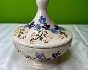 May include: A ceramic trinket box with a lid, featuring a speckled beige finish and adorned with blue and yellow floral designs. The box has a pedestal base and a pointed finial on the lid. The floral details are raised and add texture.