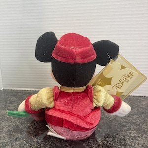 May include: A plush Mickey Mouse toy wearing a red velvet hat and matching red vest over gold sleeves. The character has black ears and a white tag attached that reads The Disney Store. The plush is viewed from the back, showing the red fabric.