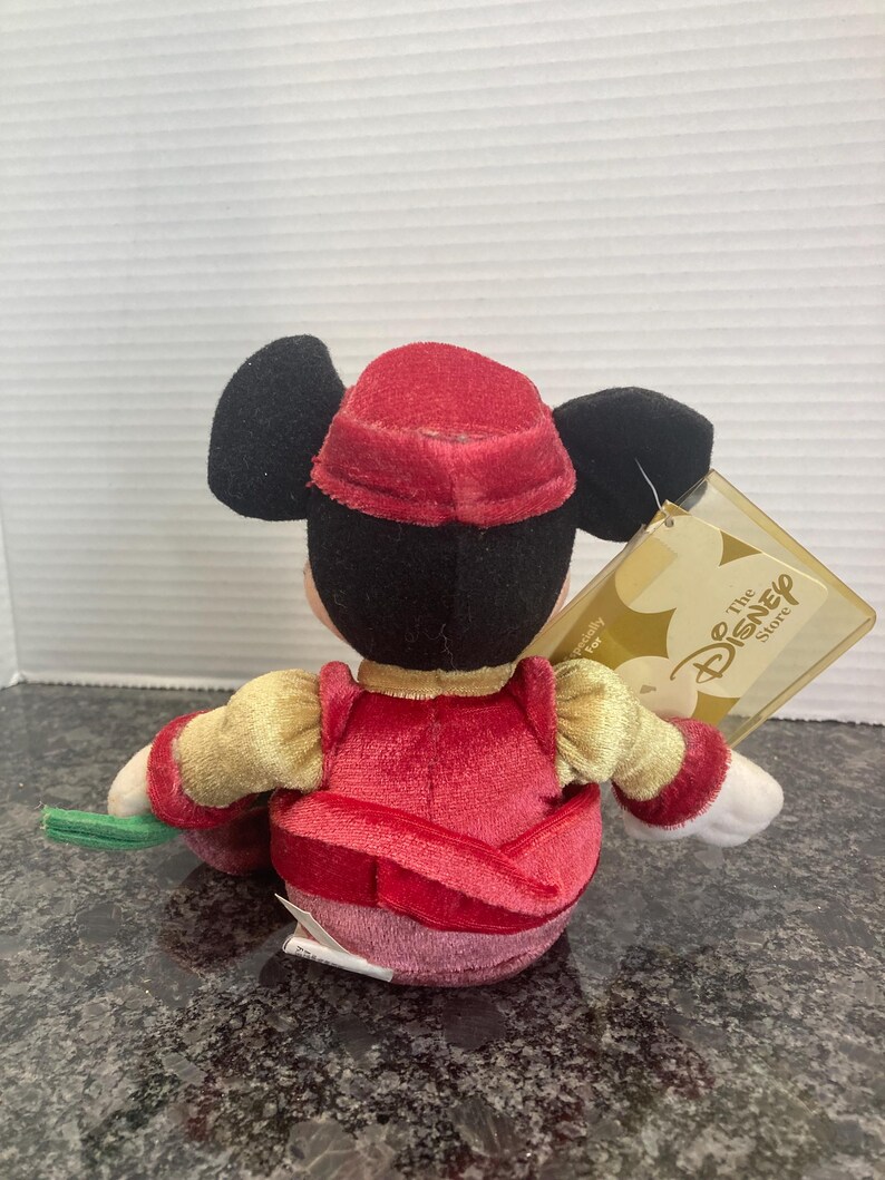 May include: A plush Mickey Mouse toy wearing a red velvet hat and matching red vest over gold sleeves. The character has black ears and a white tag attached that reads The Disney Store. The plush is viewed from the back, showing the red fabric.