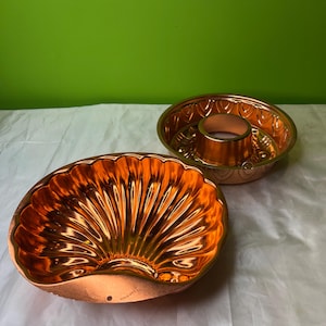 May include: Two copper-coloured bundt cake moulds. One is a fluted, shell-shaped mould. The other is a round mould with a central tube and decorative patterns around the rim. Both are made of shiny metal.