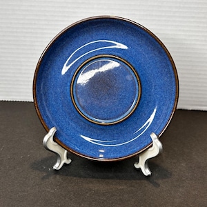 Pode incluir: Um pires de cerâmica azul com borda marrom e centro rebaixado. O pires é exibido em um suporte de metal prateado sobre um fundo branco e preto. O pires tem um acabamento brilhante.