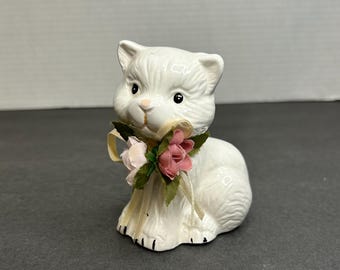 May include: A white ceramic figurine of a cat wearing a pink and green flower bow. The cat is sitting with its paws tucked under its body.