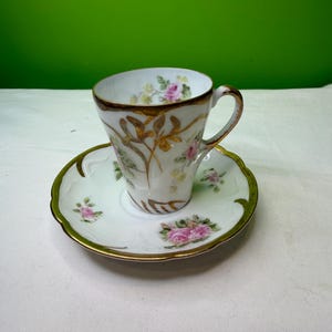 May include: White teacup and saucer set with gold trim and floral accents. The cup and saucer feature pink roses and gold leaf designs. The set is on a white surface with a green background.