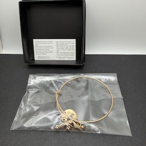 May include: A gold-tone wire bangle bracelet featuring a round charm with the word Faith and a cross charm with clear stones. The bracelet is inside a clear plastic bag, positioned in front of a black jewellery box with a white care instruction card.