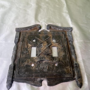 Puede incluir: Una placa de interruptor de luz vintage de metal con un diseño decorativo que presenta baquetas cruzadas, banderas y el texto "The Drum 1776".