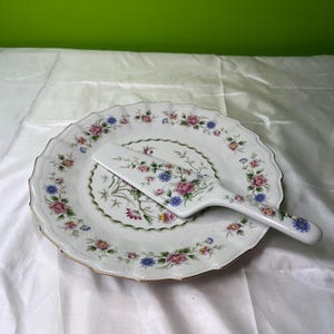 Pode incluir: Conjunto de prato e servidor de bolo em cerâmica branca. O prato tem uma borda ondulada com acabamento dourado e é decorado com padrões florais rosa, azul e amarelo. O servidor tem um design floral correspondente.