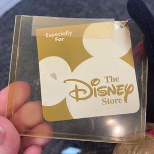 May include: A rectangular clear plastic tag holder containing a gold and white card with the text Especially For The Disney Store. The card features a large white Mickey Mouse head silhouette against a gold background with the logo printed in gold.