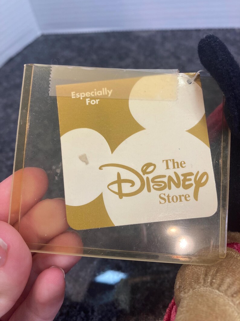 May include: A rectangular clear plastic tag holder containing a gold and white card with the text Especially For The Disney Store. The card features a large white Mickey Mouse head silhouette against a gold background with the logo printed in gold.