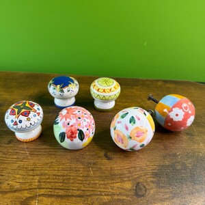 May include: A collection of seven decorative ceramic cabinet knobs. The knobs feature various colorful floral and geometric patterns, including flowers, peaches, and abstract designs. Each knob has a unique design and color scheme.
