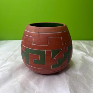 May include: A terracotta-colored ceramic vessel with a green geometric design. The vessel has a rounded shape with a dark green interior. The design is a repeating pattern of angular shapes. The background is a solid green.