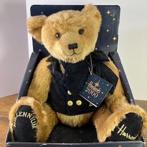 Puede incluir: Un oso de peluche Harrods Millennium de color marrón, con un chaleco negro con botones dorados y una pajarita negra. El oso está en una caja con un fondo azul marino y estrellas doradas. Las patas del oso tienen bordadas las palabras "Millennium" y "Harrods".
