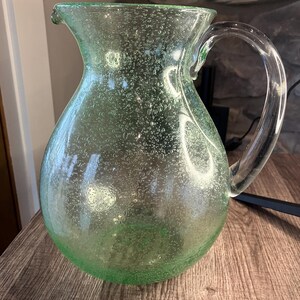 May include: A clear green glass pitcher with a textured surface and a handle. The pitcher has a round body and a wide mouth.