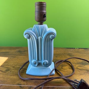 May include: A light blue ceramic lamp with a decorative, fluted design. The lamp has a brown metal socket and a brown electrical cord with a two-prong plug. The lamp is on a wooden surface with a green background.