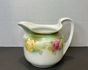 May include: A white porcelain jug with a green and white floral design. The jug has a wide mouth and a handle. The flowers are yellow, pink, and white.