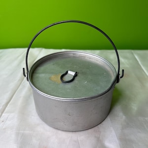 Può includere: Una piccola pentola rotonda in alluminio con coperchio e manico in filo di ferro nero. La pentola ha una finitura argentata e un design semplice, adatta per il campeggio o la cucina all'aperto. Il coperchio ha una piccola maniglia nera.
