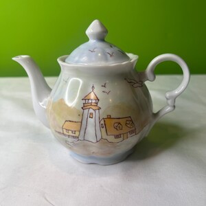 May include: A white porcelain teapot with a blue and brown lighthouse and houses painted on it. The teapot has a lid with a small knob on top and a handle on the side.