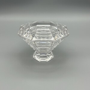 Op de afbeelding: Een heldere, facetgeslepen glazen kom met een geometrisch ontwerp. De kom heeft een brede, achthoekige bovenkant en een voetstuk. Het glas reflecteert licht. De kom wordt waarschijnlijk gebruikt voor decoratieve doeleinden of om te serveren.