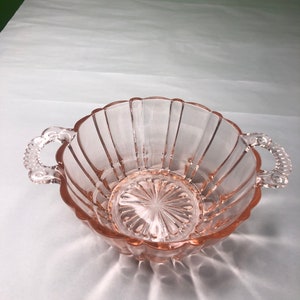 May include: A pink glass bowl with two handles. The bowl has a fluted design and a scalloped edge.