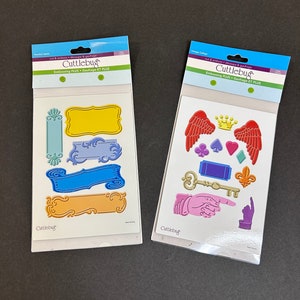 May include: Two packs of Cuttlebug embossing dies. The first pack contains dies for creating ornate frames and banners in blue, yellow, and orange. The second pack contains dies for creating wings, a crown, a key, and pointing hands in red, purple, and gold.