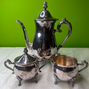 May include: A silver tea set with a tall teapot, a sugar bowl with a lid, and a creamer. The set has ornate handles and rests on small, decorative feet. The teapot has a long spout and a decorative finial on the lid.
