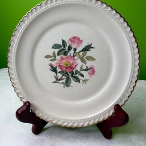 Pode incluir: Um prato de cerâmica branco com uma borda dourada e um design floral com rosas cor-de-rosa e folhas verdes. O prato tem uma borda ondulada e é exibido em um suporte marrom escuro.